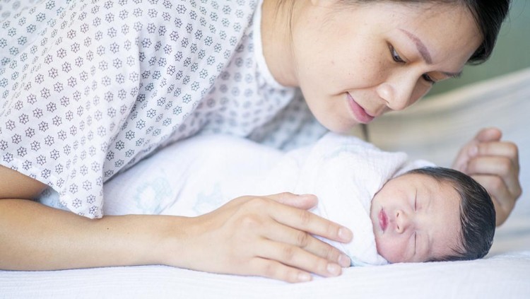 Shortly after birth a newborn baby boy is wrapped tightly in a swaddling blanket and laid on the bed to rest&period;  The Asian baby has a full head of dark hair and has his eyes closed as he sleeps peacefully&period;  His Mother is leaning in closely over him to watch as he sleeps&period;
