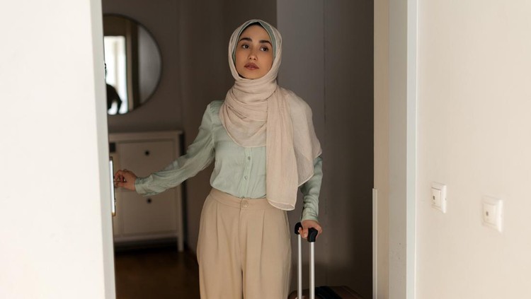 Refugee Muslim woman wearing hijab standing at the doorway holding suitcase&comma; looks worried and depressed