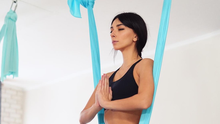 Full length of fit young woman doing antigravity yoga exercises in studio&period; Namaste butterfly pose&period; Side view