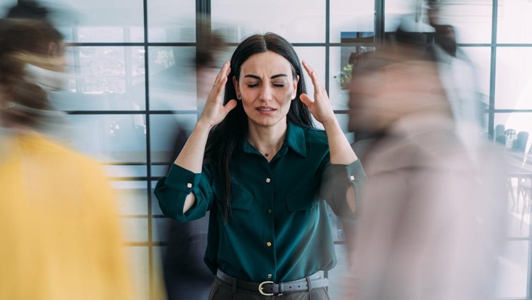 Shot of overwhelmed businesswoman holding her head while working in a chaotic office environment with blurred colleagues rushing around&period; Stressed businesswoman experiencing a headache while standing in a busy modern office environment with blurred colleagues moving around in the background&period;