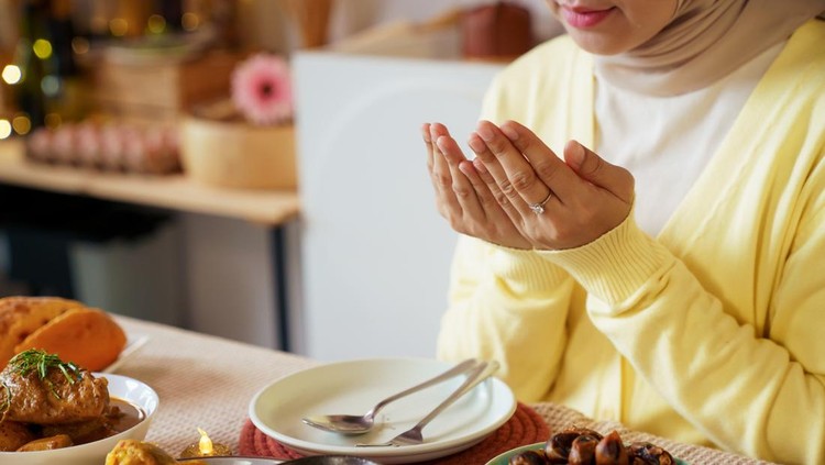 Muslim woman in hijab raises her hands in prayer before enjoying an iftar meal at home&period; Quiet ritual reflects devotion&comma; gratitude&comma; and the meaningful pause observed before breaking fast during Ramadan evening&period;