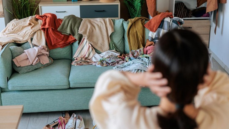 Woman standing in front of a cluttered living space filled with clothes&comma; holding her head in frustration&period; The image captures anxiety&comma; decision fatigue&comma; and home organization challenges&period;