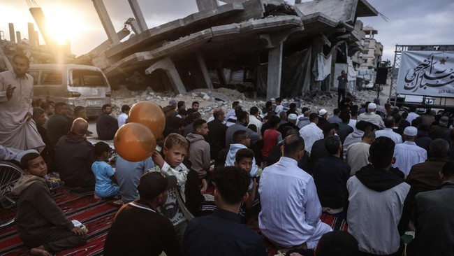 A boy holding balloons stands among Palestinians gathering on a heavily damaged street to perform morning prayers marking the start of Eid al-Adha, the Feast of Sacrifice, in Khan Yunis in the southern Gaza Strip on May 27, 2026. Since October 10, a