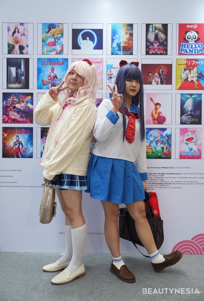 Dua cosplayer ini mengikuti gaya seragam sekolah khas Akademi Shuchiin. Cosplayer kiri dengan cardigan rajut krem untuk kesan santai, sedangkan cosplayer kanan menggunakan seragam sailor fuku dengan pita merah besar di dada.