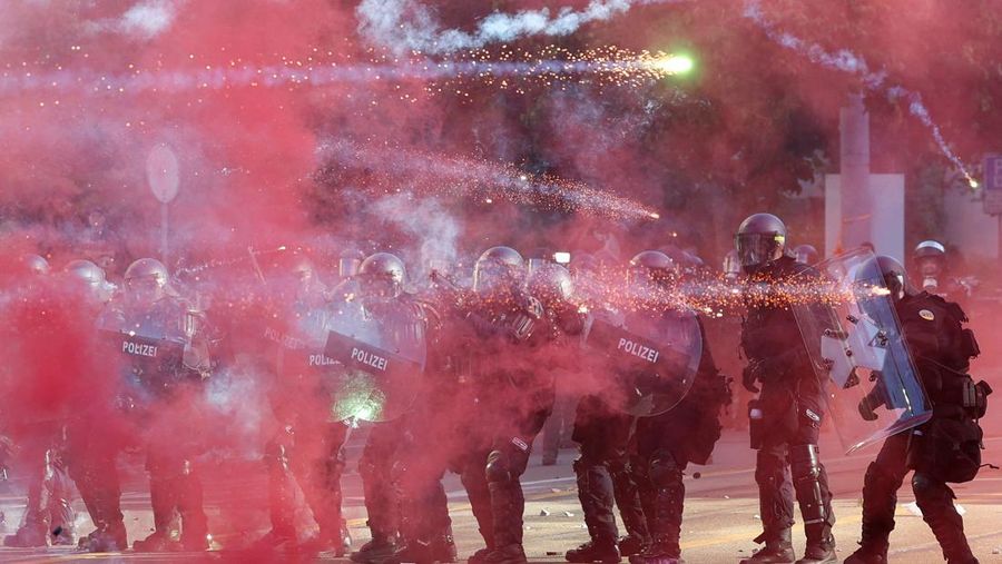 Petugas polisi anti huru hara menggunakan perisai untuk melindungi diri mereka sendiri, sementara kembang api beterbangan di udara, selama bentrok dengan demonstran, di tengah protes terhadap KTT G7 nan bakal datang di Evian-les-Bains di Prancis, di Jenewa, Swiss, 14 Juni 2026.