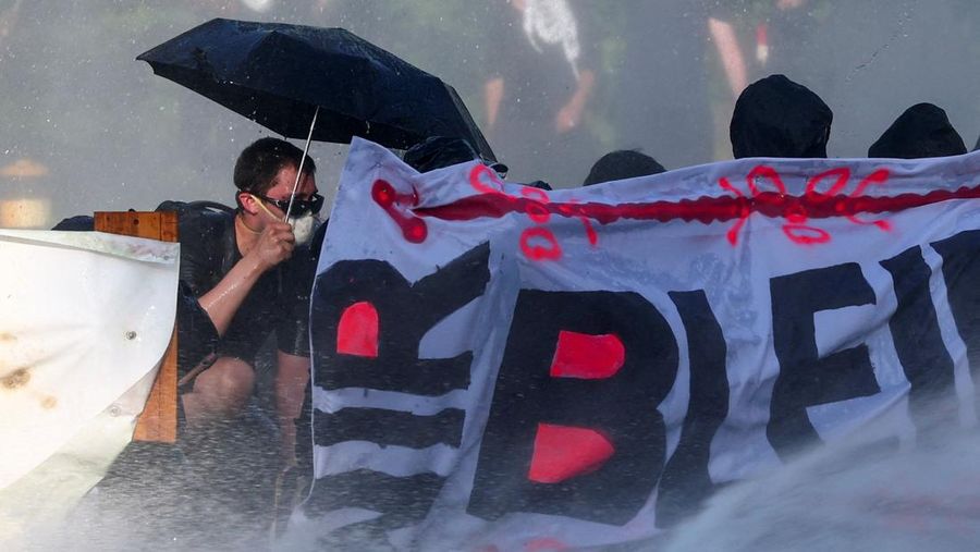 Seorang demonstran memegang payung sembari berlindung di kembali spanduk saat polisi anti huru hara menyiram mereka dengan air dari meriam air selama protes menentang KTT G7 nan bakal datang di Evian-les-Bains, Prancis, di Jenewa, Swiss, 14 Juni 2026.