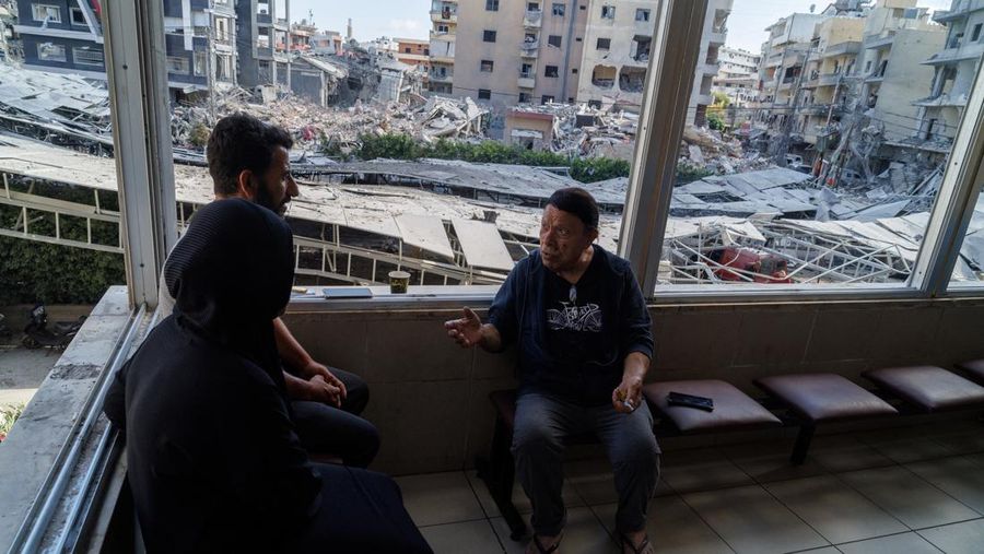 Youssef Fares, 90, whose home of more than 70 years was damaged by an Israeli airstrike, chats with fellow displaced people as they sit beside broken windows of Jabal Amel hospital, where he has taken shelter with his family, overlooking buildings da