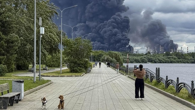 People walk in a park as black smoke rises from the area of the Russian oil producer Gazprom Neft's Moscow oil refinery on the south-eastern outskirts of Moscow on June 18, 2026. Moscow was fending off a