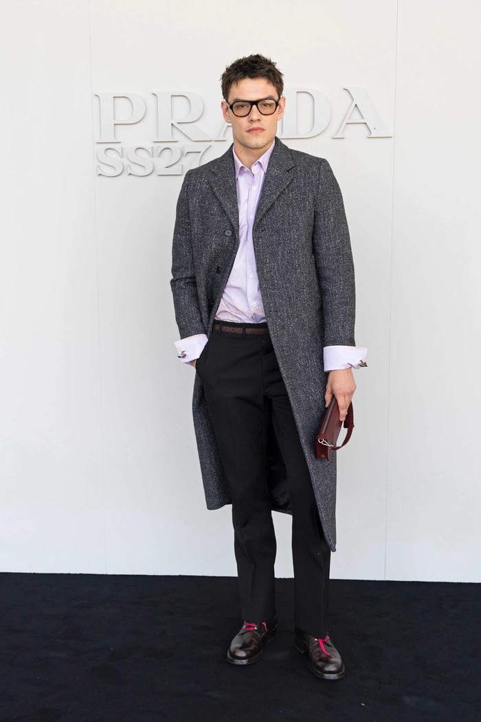 Fashion show Prada jadi momen untuk tampil posh. Louis Partridge rapi mengenakan kemeja lilac dengan tailored pants hitam, lalu ditumpuk dengan coat abu-abu berpotongan bersih. Styling dengan kacamata full frame nan retro, clutch, hingga skinny belt jadi detail yang unik yang melengkapi gayanya/ Foto: Dok. Prada