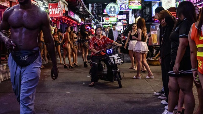 People walk past bars and clubs along a street in Pattaya on July 2, 2026. Scantily dressed and wearing stiletto heels, hundreds of girls, some apparently very young, form a seedy guard of honour under the lights of Soi 6, a street in one of Pattaya'