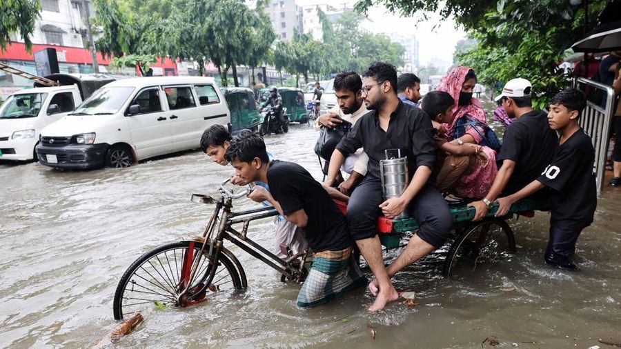 Orang-orang menyeberangi jalan nan tergenang banjir menggunakan becak setelah hujan deras di Dhaka, Bangladesh, 12 Juli 2026.
