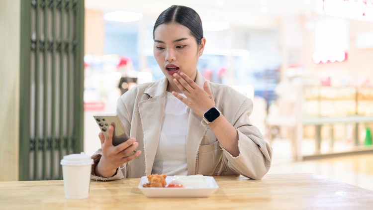 A young Southeast Asian Thai businesswoman from the Isan region looking at her smartphone with a shocked expression&comma; covering her mouth with her hand&period; She is wearing a professional beige business blazer and sitting at a wooden table in an indoor food court&period; In front of her is a white takeaway coffee cup alongside a plate of fried chicken and sticky rice&period; Horizontal framing with soft background bokeh and copy space above her&period;