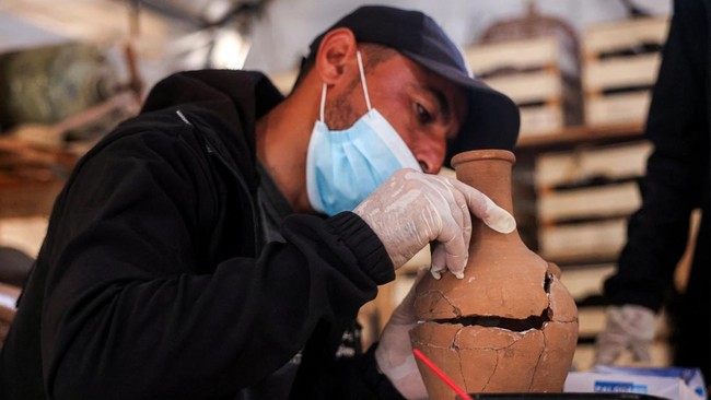 Seorang staf dari Asosiasi Budaya dan Seni Mayasem sedang menyatukan kembali sebuah potongan tembikar, sementara staf lainnya memperbaiki dan merestorasi secara ilmiah sejumlah artefak yang dikumpulkan selama perang di Gaza, di fasilitas penyimpanan