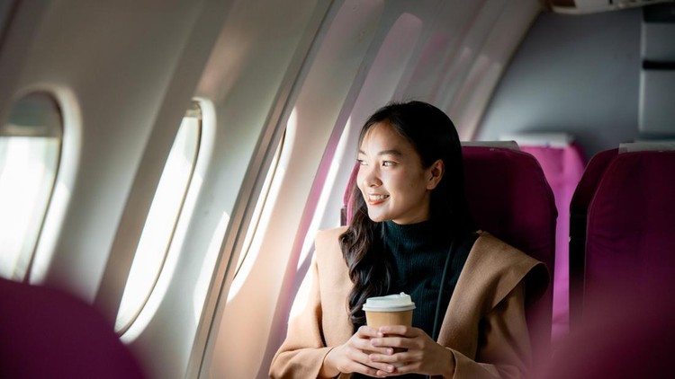 Young woman smiles while holding a coffee cup and looking out the window during a relaxing airplane flight