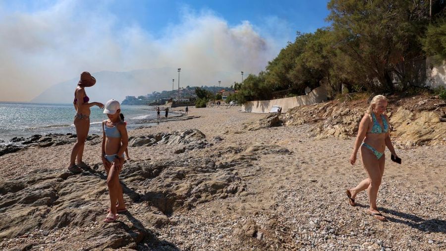 Orang-orang melangkah di pantai sementara asap mengepul dari kebakaran rimba nan terjadi di dekat desa Plomari di pulau Lesbos, Yunani, 29 Juli 2026.