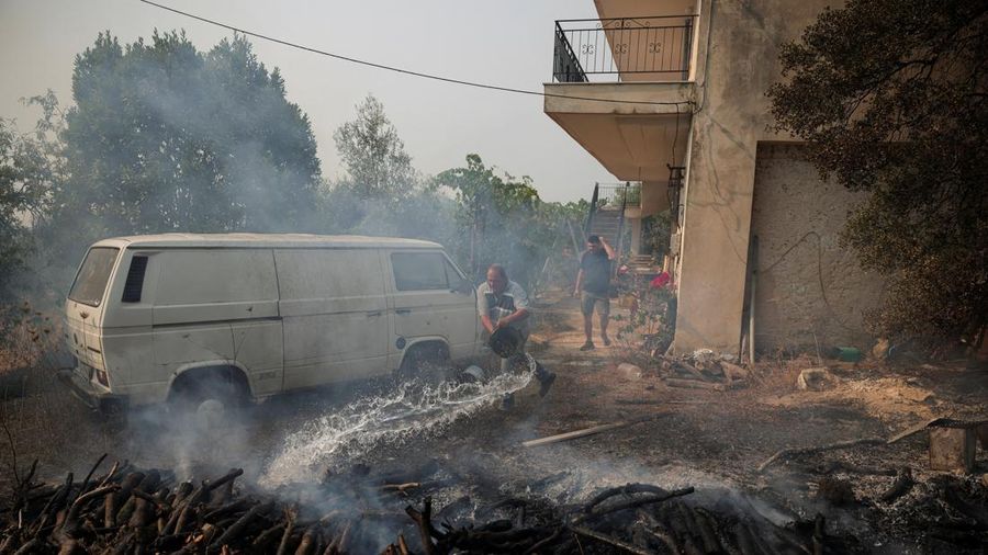 Seorang penduduk menyiramkan air ke kayu nan tetap berkepul di luar sebuah rumah setelah kebakaran rimba Boeotia melanda Veniza, Yunani, 2 Agustus 2026.