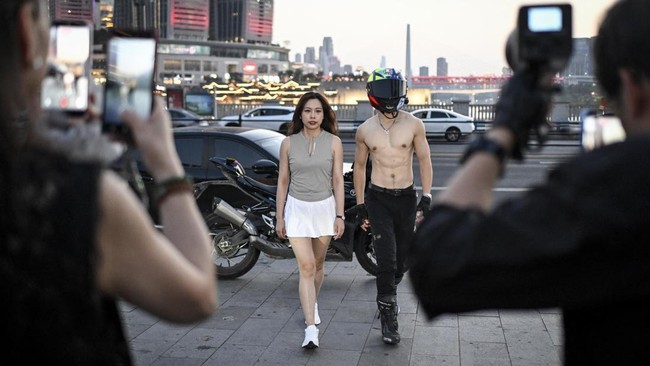 This picture taken on July 20, 2026 shows a woman posing with a motorcycle rider offering photo and video services along a street in Chongqing, southwestern China. The roar of engines fills the night air in China's Chongqing city as tourists awkwardl