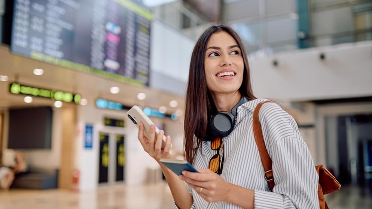 Smiling tourist holding her passport and smartphone while looking at the flight information display at the airport