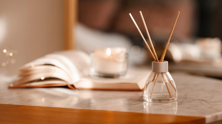 Liquid home fragrance in glass bottle with bamboo sticks staying on marble coffee table over candle and open book in room close up&period; Aromatherapy&period;