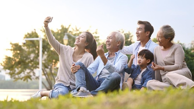 three generation happy asian family sitting on grass taking a selfie using mobile phone outdoors in park