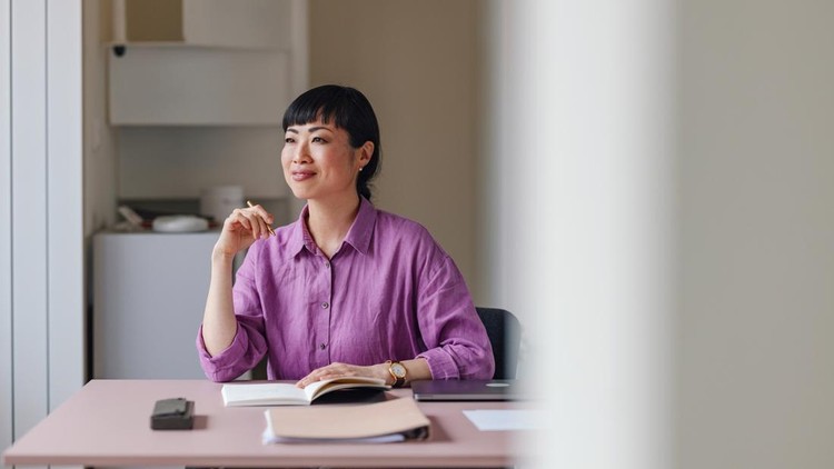 Contemplative Asian businesswoman with pen in hand sitting at desk with a notebook in a modern office&period;