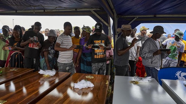 Mourners use their mobile phones as they file past coffins during a memorial service held for victims of the capsized Rural Infrastructure Development Agency (RIDA) passenger ferry at Nyamhunga Stadium in Kariba, on August 13, 2026. A government pass