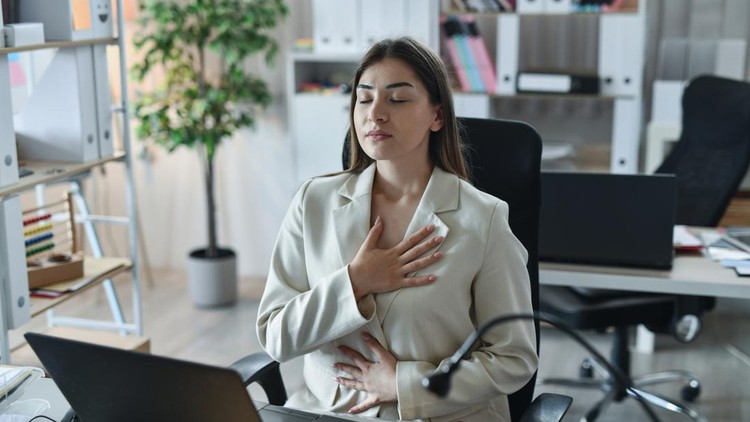 Female professional sitting at her office desk practicing deep breathing exercises&comma; calmly inhaling and exhaling to reduce stress&comma; anxiety and mental fatigue during a busy workday&period;