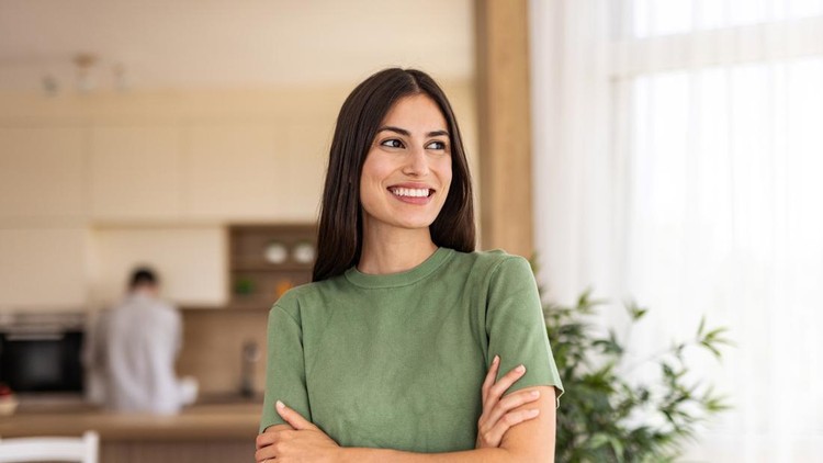Portrait of a cheerful young woman standing with arms crossed in a modern kitchen&comma; smiling and looking away&comma; savoring the joys of domestic life and the comfort of her home