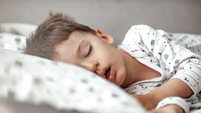 Little blond boy sleeping in his bed. Restless sleep of a baby with a cold. The kid sleeps with his mouth open. My little boy sleeping by the light of the window.