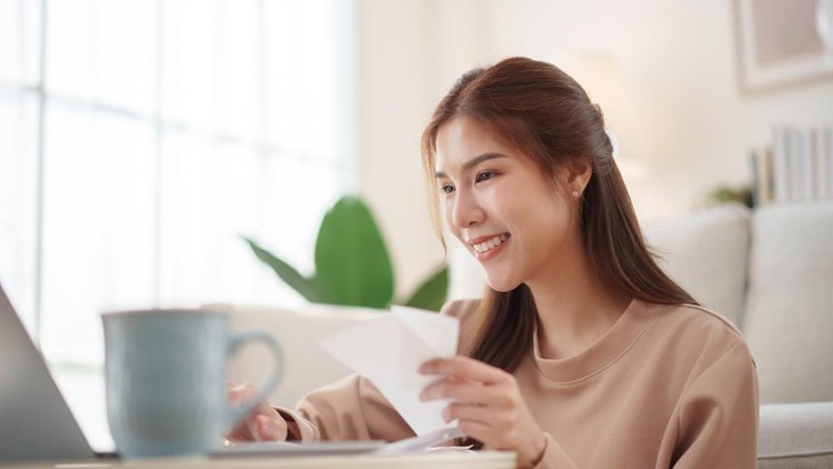 Cheerful young Asian woman holding receipts and working on a laptop in a living room&comma; sitting at a modern table&comma; organizing financial documents in a relaxed home&period;