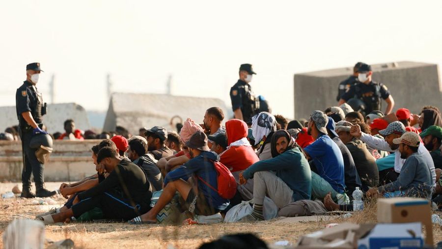 Para migran berkumpul di dekat sebuah kamp darurat di tepi pantai untuk dipindahkan ke tempat penampungan resmi di beragam letak di kota tersebut setelah pihak berkuasa membersihkan area itu—menyusul penyeberangan massal dari Maroko—di wilayah kantong Spanyol di Afrika Utara, Ceuta, pada 20 Agustus 2026.