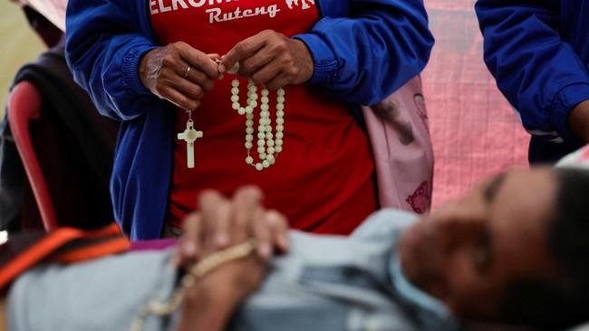 A patient receiving treatment and a relative standing next to them, both pray hold rosaries, in a makeshift tent outside the Ben Mboi Manggarai regional hospital in the aftermath of an earthquake, in Ruteng, Manggarai regency, East Nusa Tenggara prov