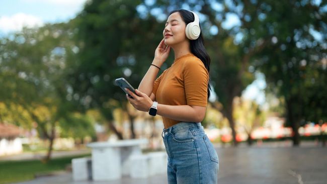 Young adult woman wearing wireless headphones, listening to music on a smartphone, relaxing outdoors