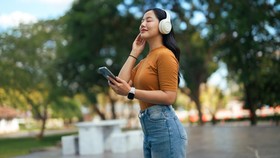 Young adult woman wearing wireless headphones, listening to music on a smartphone, relaxing outdoors