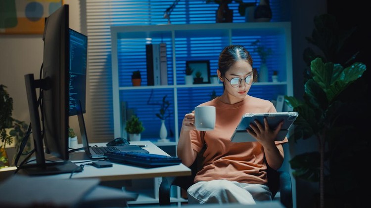 Gen Z Asian computer programmer drinks coffee while read digital tablet in home office at night&period; Taking a relaxing coffee break&comma; freelance work&comma; software development&comma; artificial intelligence&period;