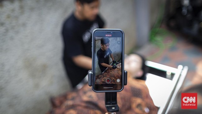 Di sebuah bangunan di kota Bandung, Yoka Brahma Putra menjual kain kafan sambil berseru checkout sebelum meninggal, ditemani sosok berbalut kain kafan mirip pocong yang berdiri diam di belakangnya, dan siaran itu viral. (CNN Indonesia/ Adhi Wicaksono