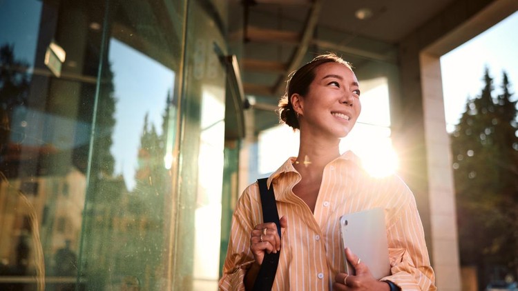 Young Asian businesswoman walking confidently near an office building&comma; holding a tablet and smiling while looking away&comma; beautifully backlit by the warm glow of the setting sun