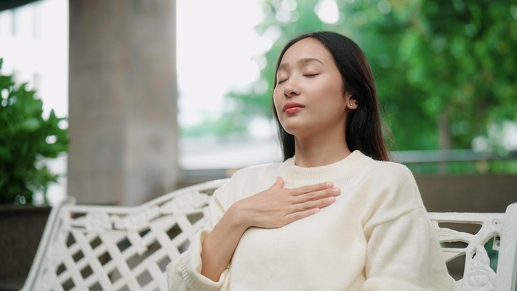 Woman gently resting one hand on her chest and focusing on the breath while sitting on a bench in the garden&period;