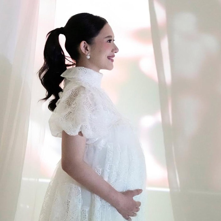<p>Tema pertama maternity shoot, Athina tampil cantik dan anggun mengenakan dress putih dengan kerah tinggi dan potongan A-line serta ruffle. Penampilannya disempurnakan dengan flat shoes, serta asesoris kalung, gelang dan anting. (Foto: @rharyantoracing)</p>