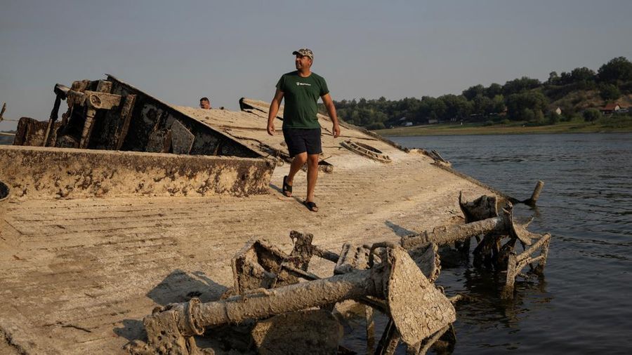 Seorang laki-laki berdiri di atas kapal perang Jerman dari Perang Dunia II nan tenggelam dan sekarang muncul ke permukaan akibat surutnya air Sungai Danube—yang disebabkan oleh kekeringan dan suhu panas ekstrem—di dekat Prahovo, Serbia, pada 13 Agustus 2026.