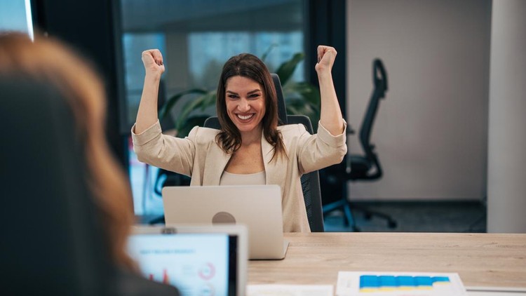 Smiling businesswoman raising fists in celebration&comma; achieving success while working on a laptop at her office desk&comma; expressing happiness and accomplishment from a business victory or goal