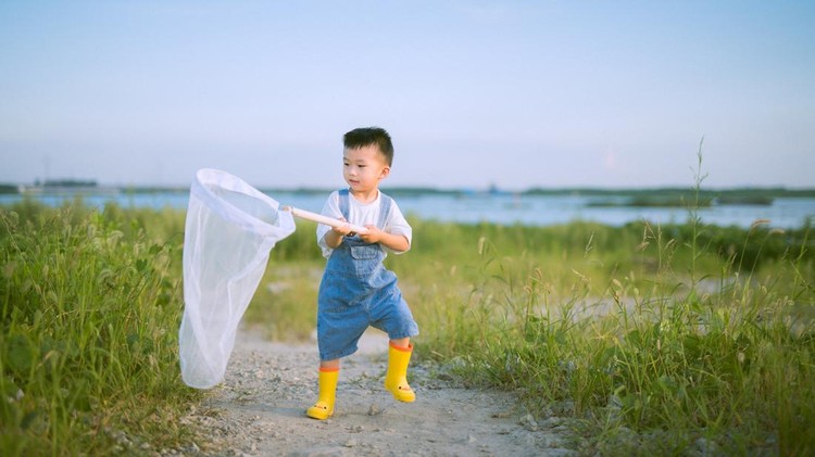 A cheerful young Asian boy dressed in blue overalls and yellow boots holds a white butterfly net while standing on a grassy path by a lake&comma; enjoying a sunny day outdoors&period; The bright blue sky and natural surroundings enhance the playful and adventurous atmosphere of the scene&period;