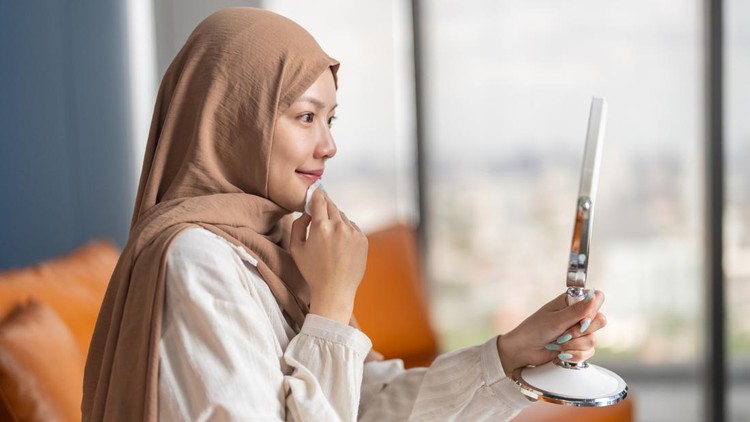 Beautiful Asian Muslim woman wearing hijab using cotton pad for self care and wellness&period; Happy female looking in mirror enjoying daily beauty care and healthy facial skincare routine at home
