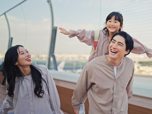 A family is walking outdoors near an airport area. A father is carrying a young child on his shoulders while the child stretches their arms outward. The mother is walking beside them, smiling. Airport infrastructure and aircraft are visible in the ba
