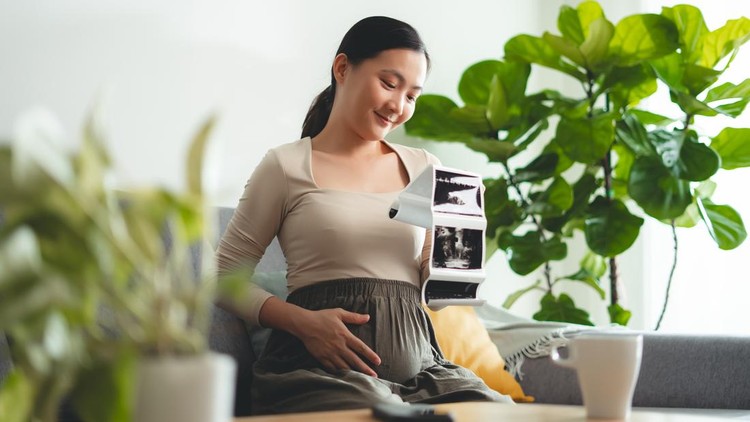 Beautiful young pregnant Indonesian woman looking at ultrasound pictures while sitting on the sofa in the living room&period;