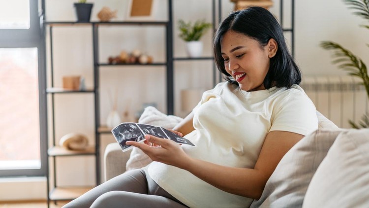 Beautiful young pregnant Indonesian woman looking at ultrasound pictures while sitting on the sofa in the living room&period;