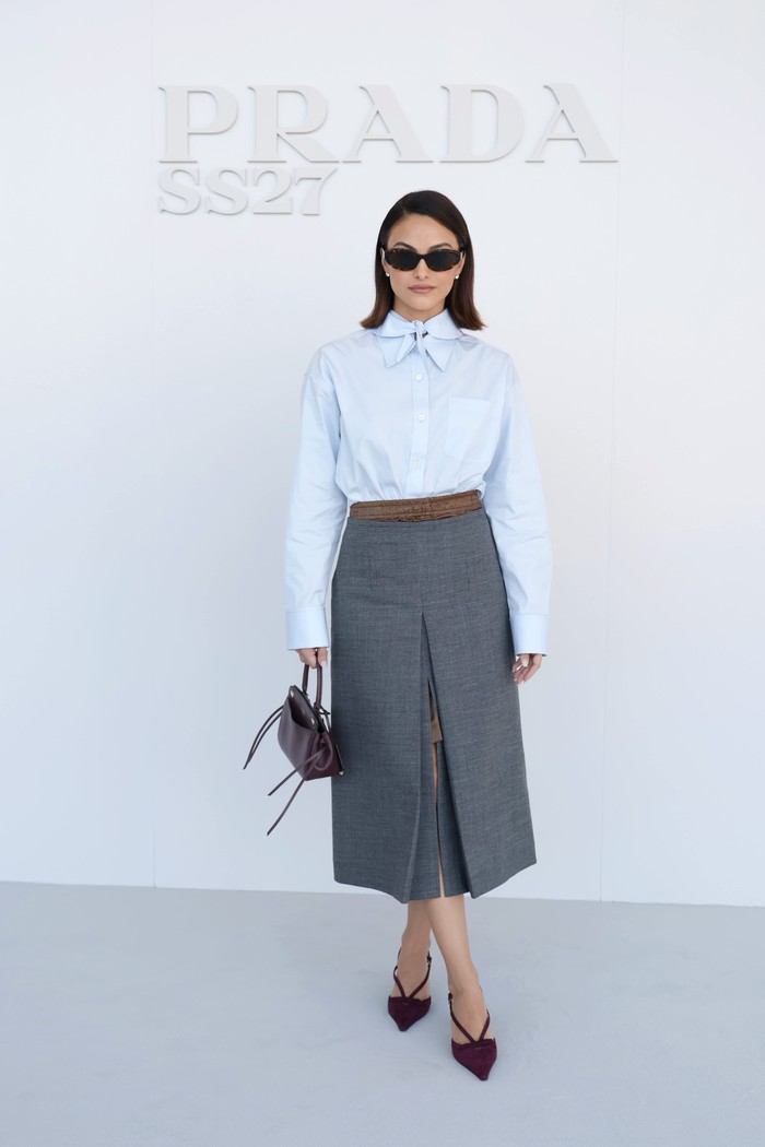 Camila Mendes memadukan midi skirt abu-abunya dengan kemeja biru muda. Detail yang quirky, seperti kerah pita ganda dan lapisan rok beda warna jadi daya tarik penampilannya./ Foto: Courtesy of Prada