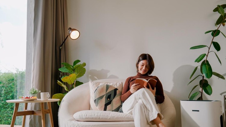 A young Asian woman turning on an air purifier in the living room before sitting on an armchair and reading&period;