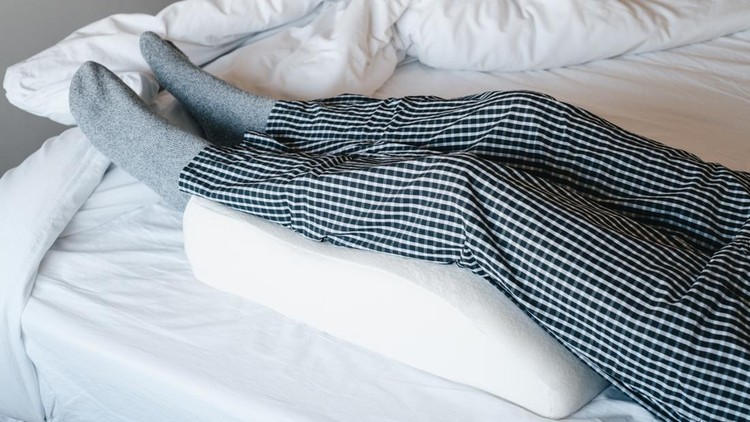 a man in bed leans his legs on a leg elevation pillow made of memory foam