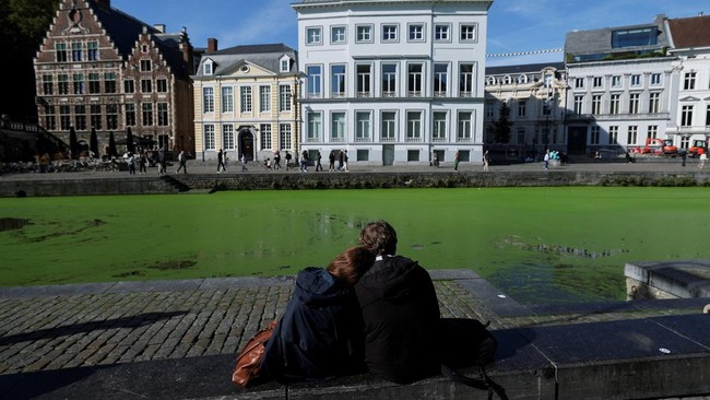 A couple sits on the bank along a duckweed-covered section of the River Lys in Ghent, Belgium, October 5, 2026. Prolonged hot and dry weather led to a surge in duckweed growth on the river following one of Europe's driest summers on record. REUTERS/S